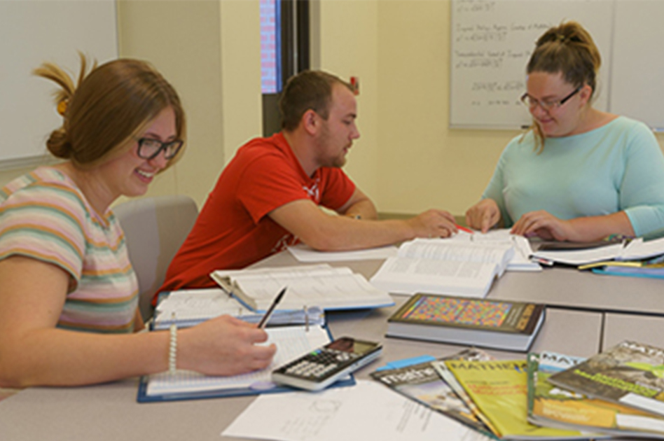 Three students sit at a table filled with math books and calculators, with the center student pointing out something in an open textbook before them. 