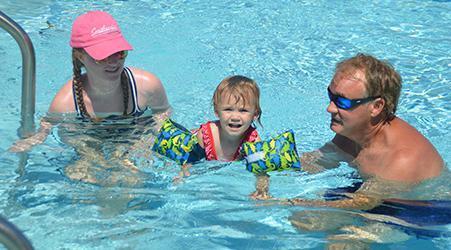 A toddler wearing arm floaties swims between two adults in a pool