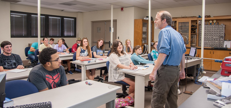 Students sitting at rows of tables in a classroom listen to an instructor as he speaks at the front of the room.