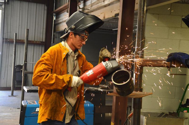 A Chipola Workforce Development student wearing a welding mask, sends sparks flying while using a grinder on a metal pipe.