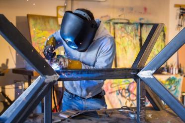 A welder wearing a welding mask and work gloves sends sparkes flying while working on the joint of a large frame. 