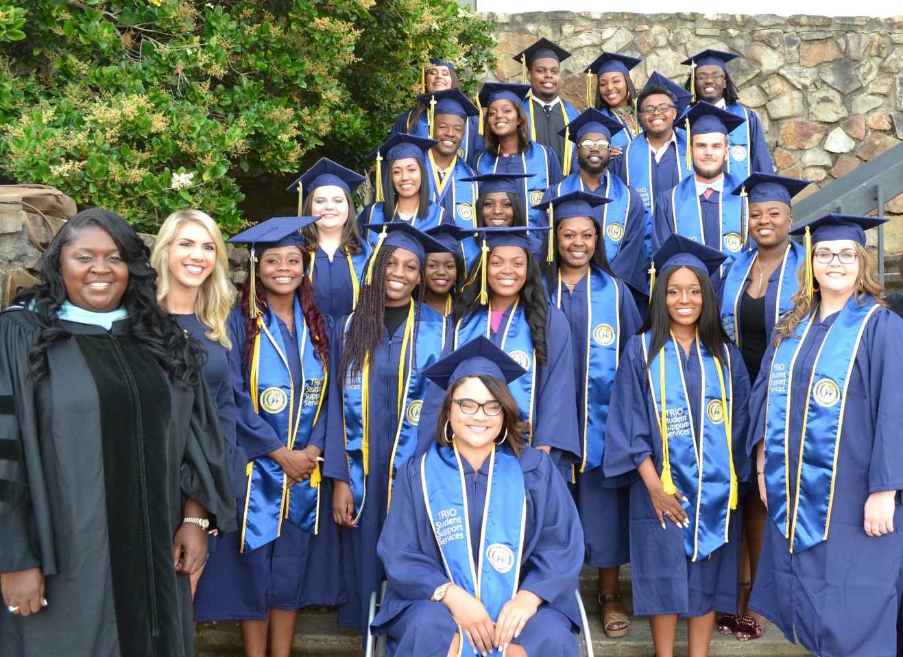 A group of 24 people pose in graduation regalia, including students wearing stoles that say 