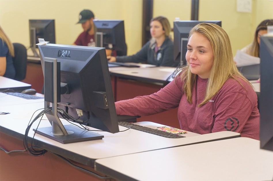 A half-similing, blonde student works at a computer in the foreground, while the bacground shows other students at rows of computers in soft focus.