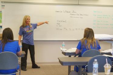 An instructor with a stethoscope around her neck stands in front of a class points to information on the white board. 