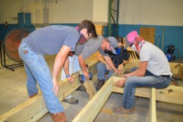 Four people wearing masks gather around a wooden frame. One holds the frame near a joint, and another holds a drill against a loose board.