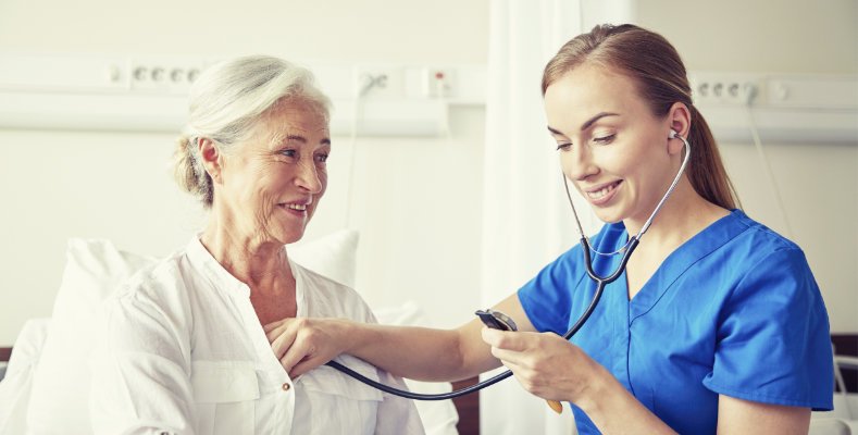 A woman in blue scrubs listens to a patient's heart with a stethoscope.