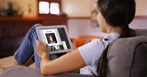 A woman sits on a couch, and a view over her shoulder shows her viewing media on a tablet propped against her knee. 