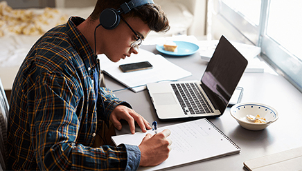 A student wearing headphones sits at a counter with a laptop and bowl of food as he writes in a notebook.