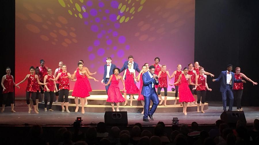 Two dozen students on stage during a show choir performance, the women in red sequined dresses and the men in suits with red bowties.